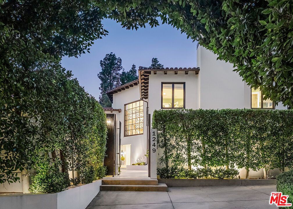 This elegant Mediterranean-style home features a crisp white stucco facade and a classic terracotta tile roof. A prominent, multi-paned window adds architectural interest, while a lush, manicured hedge provides privacy and frames the inviting entryway. The scene is captured at twilight, with warm exterior lighting highlighting the home's clean lines and the address number displayed vertically on a pillar.