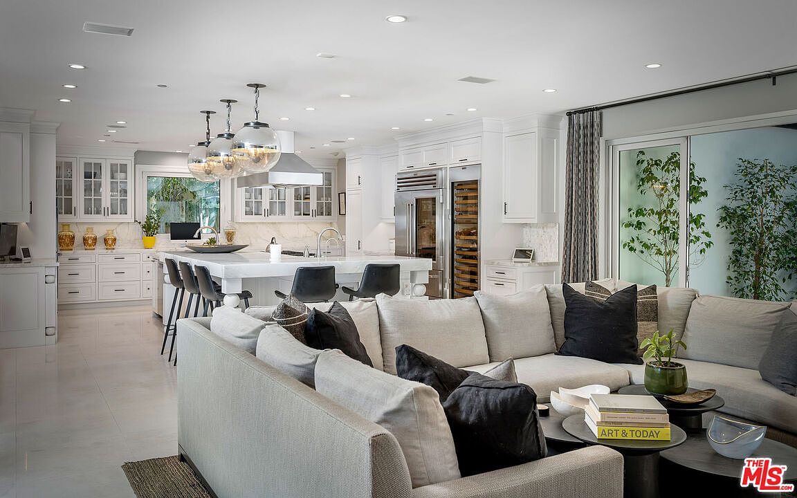 This open-concept living space seamlessly integrates a modern kitchen with a comfortable lounge area, creating an ideal layout for entertaining. The kitchen features a large central island with bar seating, white cabinetry, and high-end stainless steel appliances, while the adjacent living area is anchored by a plush, light-colored sectional sofa. Large sliding glass doors provide a view of the outdoor greenery, filling the room with natural light and enhancing the airy, sophisticated atmosphere.
