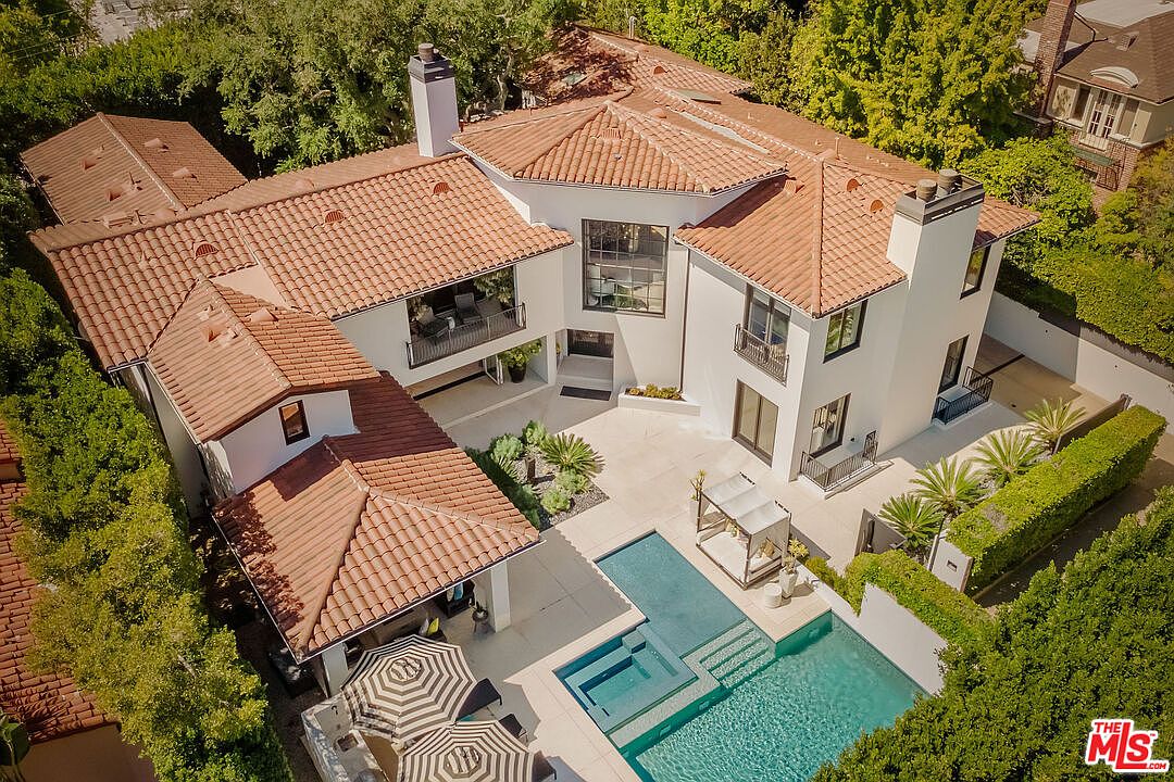 This high-angle aerial view captures a luxurious Mediterranean-style estate featuring a light-colored stucco exterior and a classic terracotta tile roof. The property includes a private swimming pool with an adjacent lounge area, covered patio space, and lush, manicured landscaping that provides significant privacy. The layout emphasizes seamless indoor-outdoor living, with multiple balconies and large glass doors connecting the interior rooms to the central courtyard.