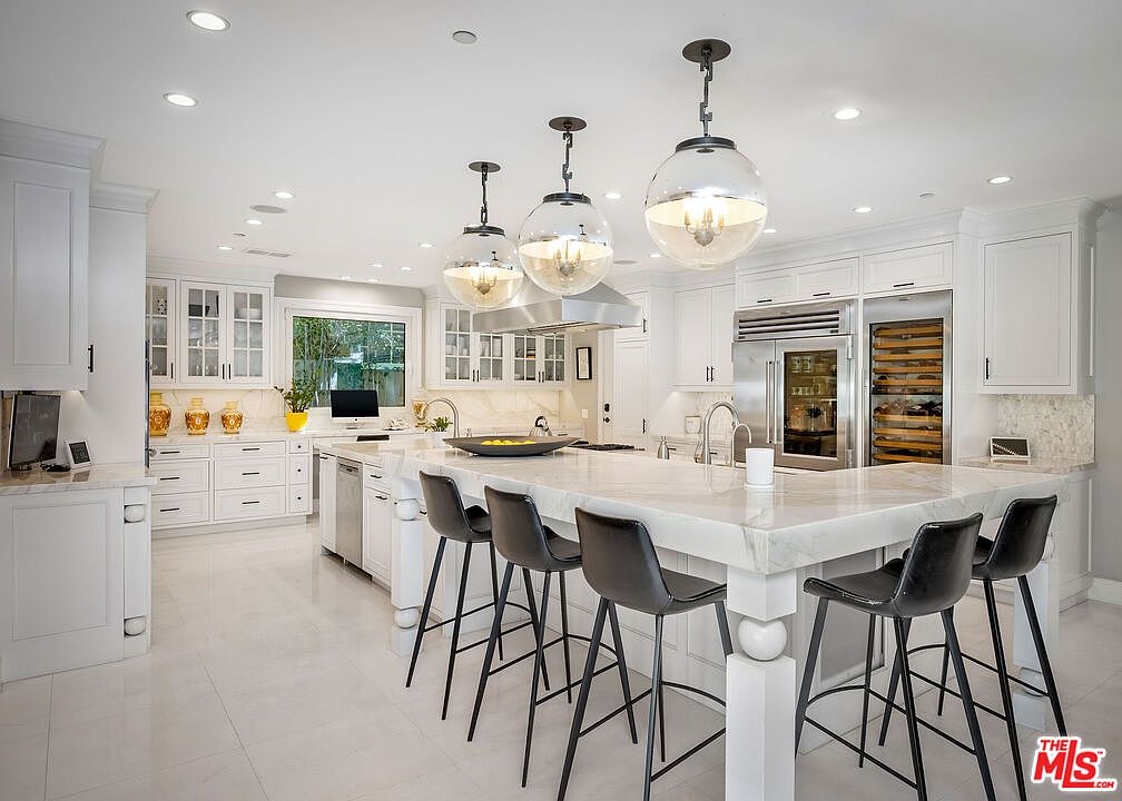This bright and spacious kitchen features white cabinetry, a large central island with stone countertops, and four dark bar stools. Three large glass pendant lights hang above the island, complementing the recessed lighting throughout the room. The space is equipped with high-end stainless steel appliances, including a built-in wine refrigerator, and offers a clean, modern aesthetic.