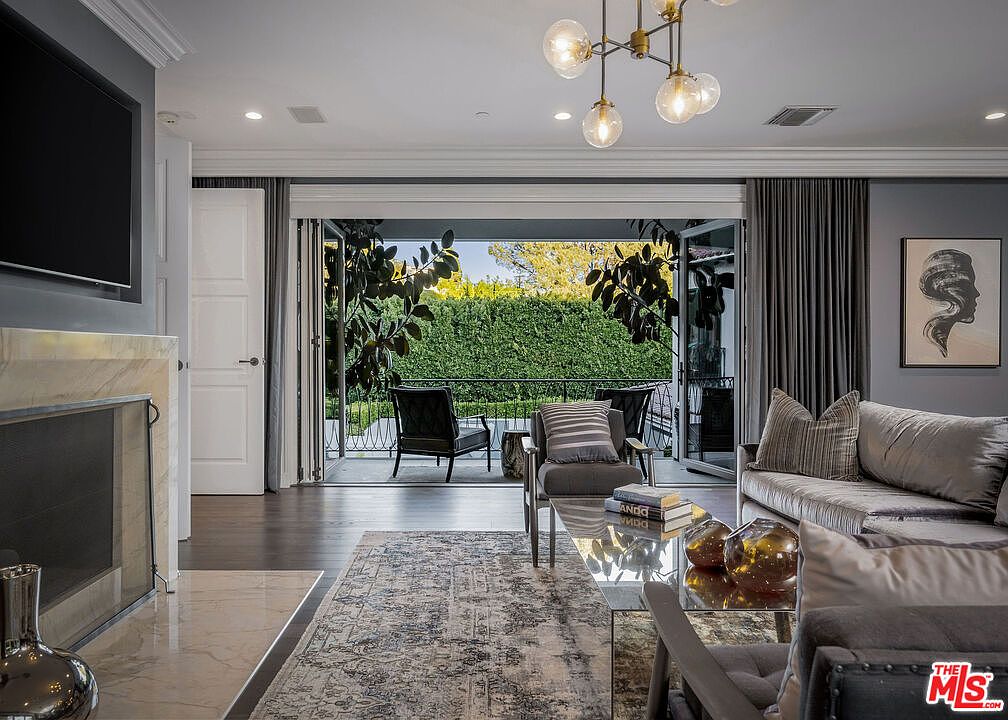 This sophisticated living room features a neutral color palette with plush seating, a patterned area rug, and a glass-topped coffee table. A stone-clad fireplace surround anchors the left side of the room, while large, folding glass doors open completely to a balcony, seamlessly blending indoor and outdoor living spaces. The room is illuminated by a modern, multi-globe chandelier, creating an inviting and airy atmosphere.