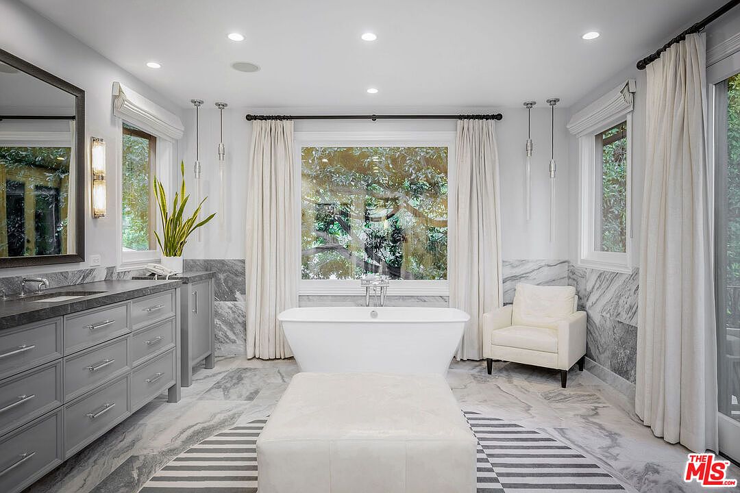 This luxurious primary bathroom features a central freestanding soaking tub positioned in front of a large window with views of lush greenery. The space is anchored by a double vanity with stone countertops and grey cabinetry, complemented by patterned stone wall accents and flooring. A plush armchair and a central ottoman add a sense of comfort and elegance to the bright, well-lit room.