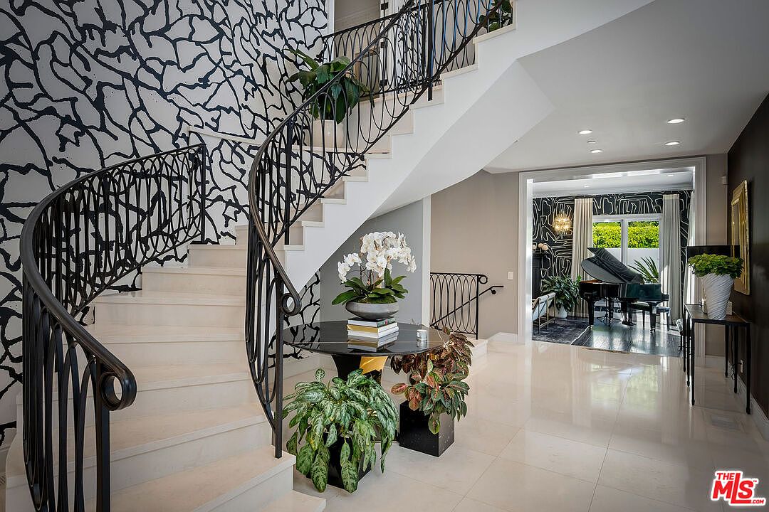 This grand foyer features a dramatic curved staircase with an intricate black metal railing set against a bold, abstract patterned wallpaper. A polished dark circular table sits at the base of the stairs, topped with a large orchid arrangement, while the glossy light-colored flooring reflects the bright interior lighting. Through the open doorway, a glimpse of a formal living area with a grand piano and matching wallpaper creates a cohesive and sophisticated aesthetic.