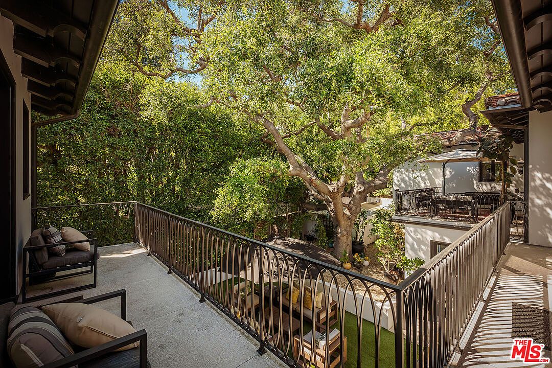 This elevated balcony offers a serene, tree-lined view overlooking a private courtyard below. The space is furnished with comfortable outdoor seating and features a decorative metal railing that provides a clear perspective of the lush greenery and the lower patio area. The scene captures a peaceful, shaded outdoor living environment perfect for relaxation.