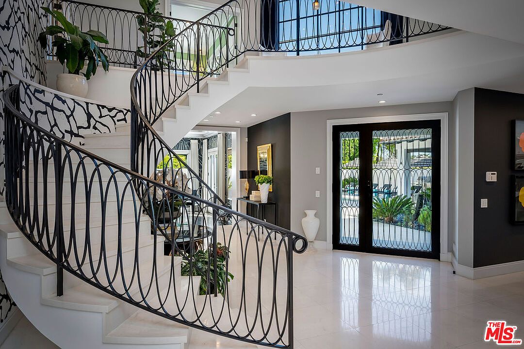 This grand foyer features a sweeping, curved staircase with intricate metal railings that serve as a focal point. The space is finished with polished light-colored flooring and high-contrast black and white wall accents, creating a sophisticated and modern aesthetic. A large double-door entry with glass panels allows natural light to flood the area, providing a clear view of the outdoor greenery.