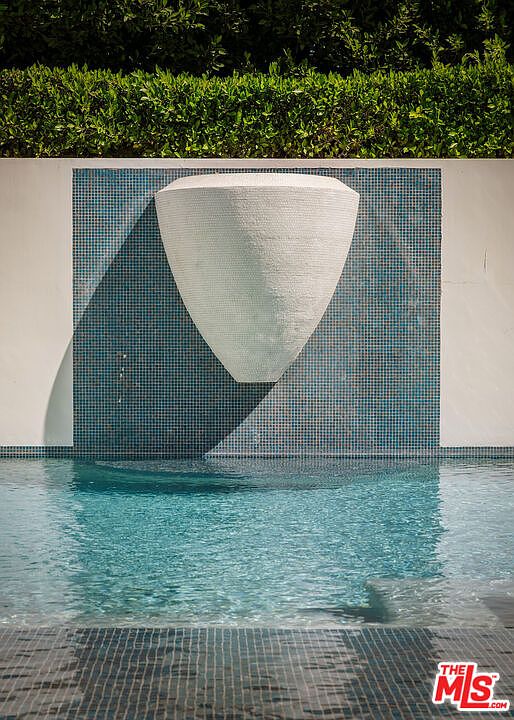 This eye-level shot captures a modern swimming pool feature, centered around a large, white, sculptural vessel mounted against a wall of blue mosaic tiles. The water cascades from the vessel into the pool below, creating a serene and artistic focal point. Lush green hedges frame the top of the wall, providing a natural contrast to the geometric lines of the water feature.