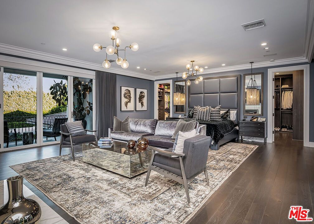 This expansive primary bedroom features a sophisticated, modern aesthetic with a large, tufted grey headboard as the focal point. The room includes a comfortable seating area with a velvet sofa and mirrored coffee table, all set upon a large, patterned area rug over dark wood flooring. Large glass doors provide access to an outdoor space, while two walk-in closets flank the bed, creating a luxurious and well-appointed suite.