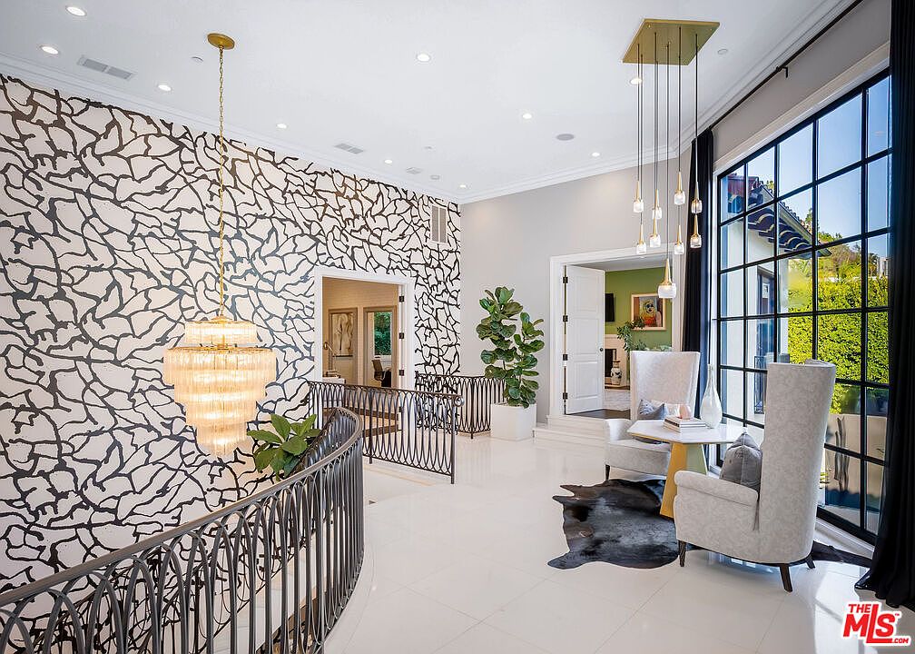 This elegant landing area features a striking accent wall with a bold, abstract black-and-white pattern that contrasts with the polished light-colored flooring. A curved metal railing overlooks the space below, while a seating area with two high-backed chairs and a small table sits beside large, grid-paned windows. The room is illuminated by a tiered crystal chandelier and a modern multi-pendant light fixture, creating a sophisticated and airy atmosphere.