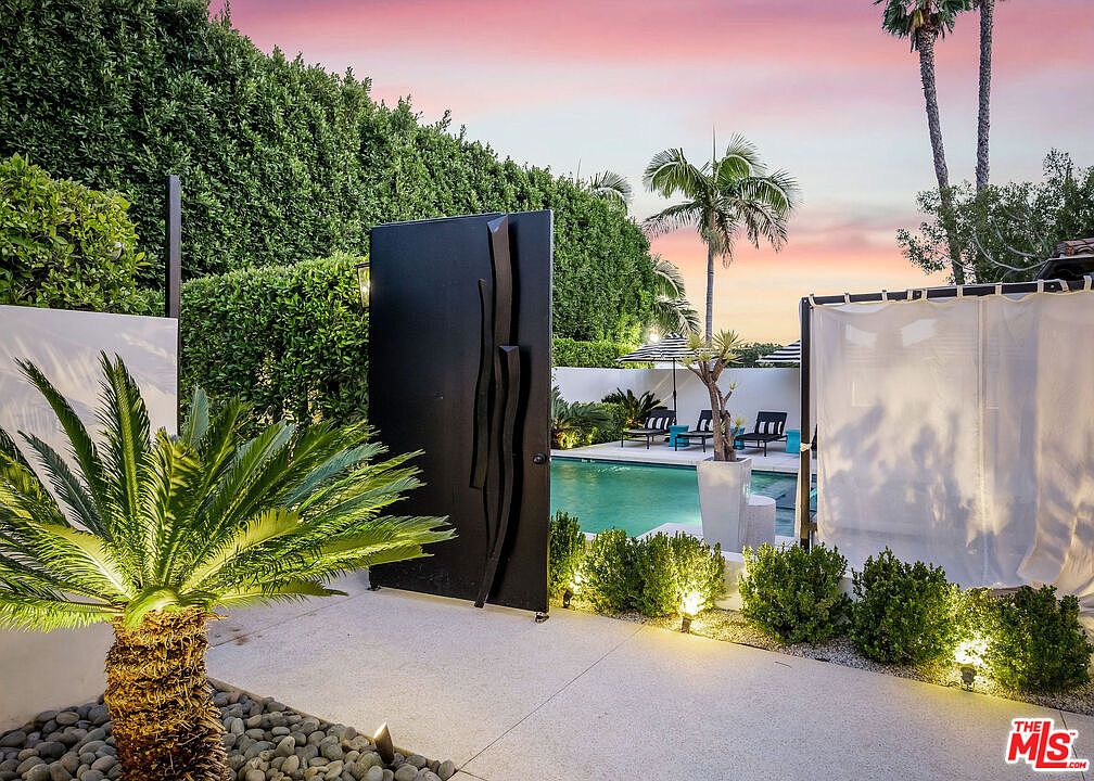 This outdoor space features a serene swimming pool framed by lush, tall hedges and a modern black metal gate. The foreground includes a manicured palm tree and landscape lighting, while the pool area is furnished with lounge chairs and umbrellas. The scene is captured at sunset, casting a warm, inviting glow over the entire backyard retreat.