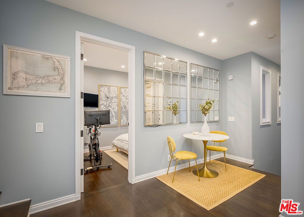 This bright hallway features light blue walls and dark wood flooring, creating a clean and inviting transition space. To the right, a small dining nook is defined by a textured rug, a round pedestal table, and two yellow chairs, accented by large grid-patterned mirrors on the wall. A doorway on the left leads into a home gym, providing a glimpse of exercise equipment and framed artwork.