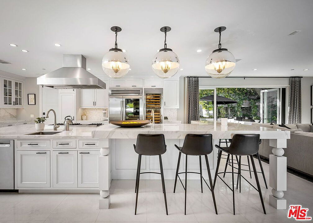 This bright, modern kitchen features a large central island with stone countertops and three black bar stools, illuminated by three spherical pendant lights. The space is equipped with white cabinetry, a stainless steel range hood, and a built-in wine refrigerator, creating a clean and sophisticated aesthetic. Large glass doors in the background provide a seamless transition to an outdoor patio area, enhancing the open-concept feel of the home.