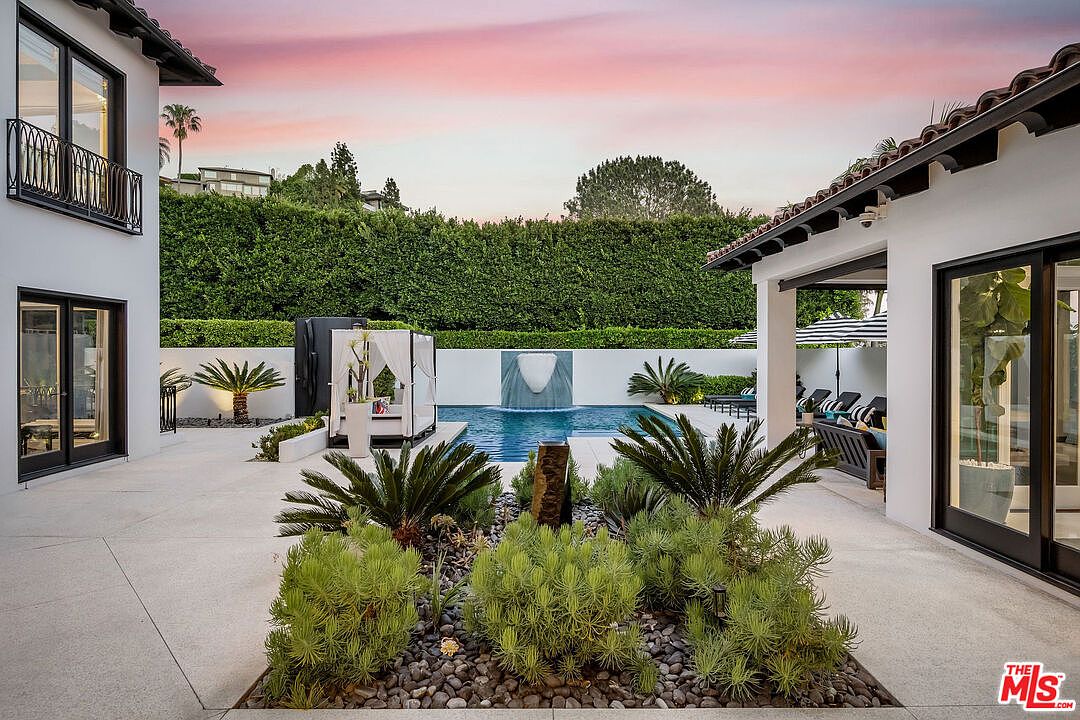 This luxurious backyard features a pristine swimming pool with a central water feature, framed by manicured hedges and a modern white-walled perimeter. A covered patio area with outdoor seating and a poolside cabana create an inviting space for relaxation and entertainment. The scene is captured at dusk, with a soft, warm sunset sky providing a serene backdrop to the contemporary landscaping and architectural design.