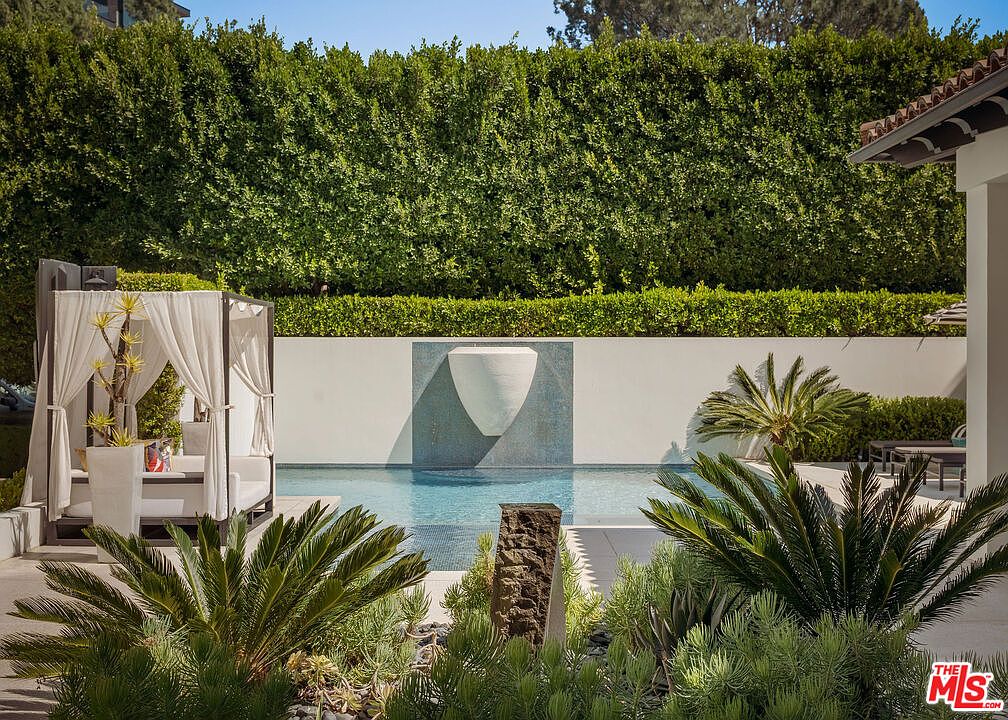 This serene outdoor space features a rectangular swimming pool centered against a tall, manicured hedge wall. A modern cabana with white drapery sits to the left, while a sculptural water feature is mounted on the rear wall above the water. The foreground is landscaped with lush cycad plants and a stone pillar, creating a private and tranquil resort-like atmosphere.