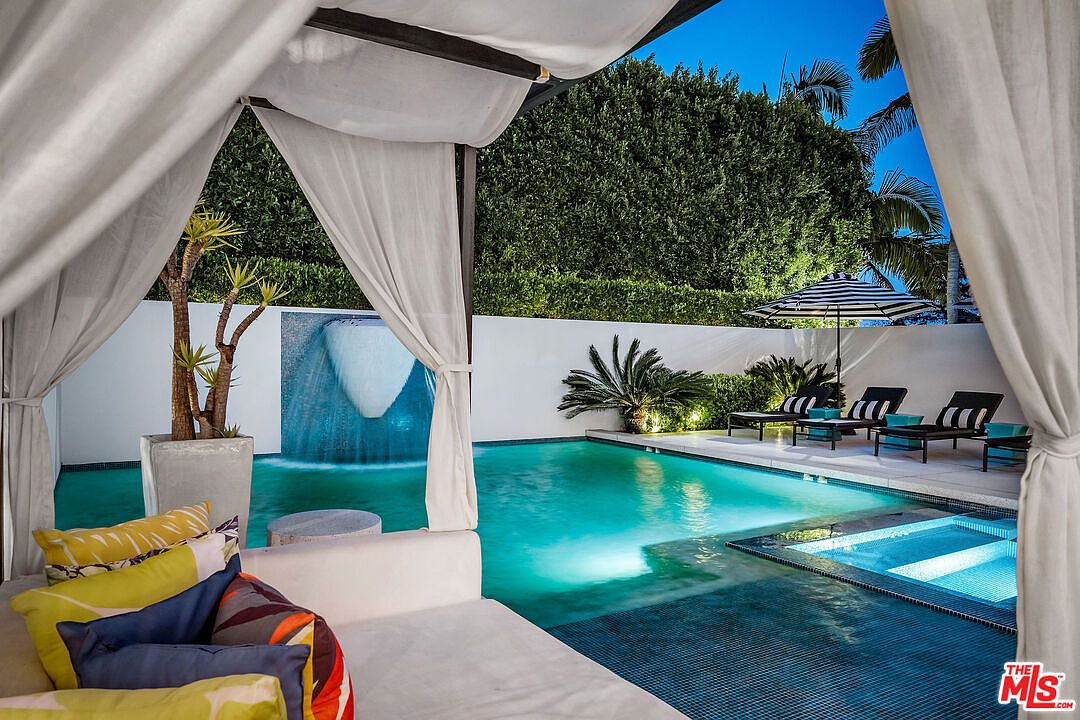 This luxurious outdoor living space features a pristine swimming pool with a built-in spa and a dramatic water feature cascading down a white wall. In the foreground, a draped cabana offers a shaded lounge area with vibrant, patterned cushions, while a row of sleek black lounge chairs sits on the adjacent patio under a striped umbrella. The scene is captured from the perspective of someone relaxing within the cabana, looking out over the illuminated pool area at twilight.