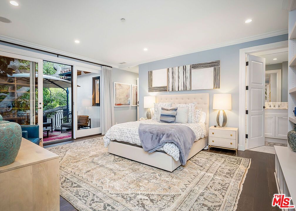 This primary bedroom features a neutral color palette with soft blue walls and a large, plush bed as the focal point. Large glass sliding doors provide seamless access to an outdoor balcony, allowing for abundant natural light and a view of the surrounding greenery. The room is anchored by a large, intricately patterned area rug over dark wood floors, creating a sophisticated and tranquil atmosphere.