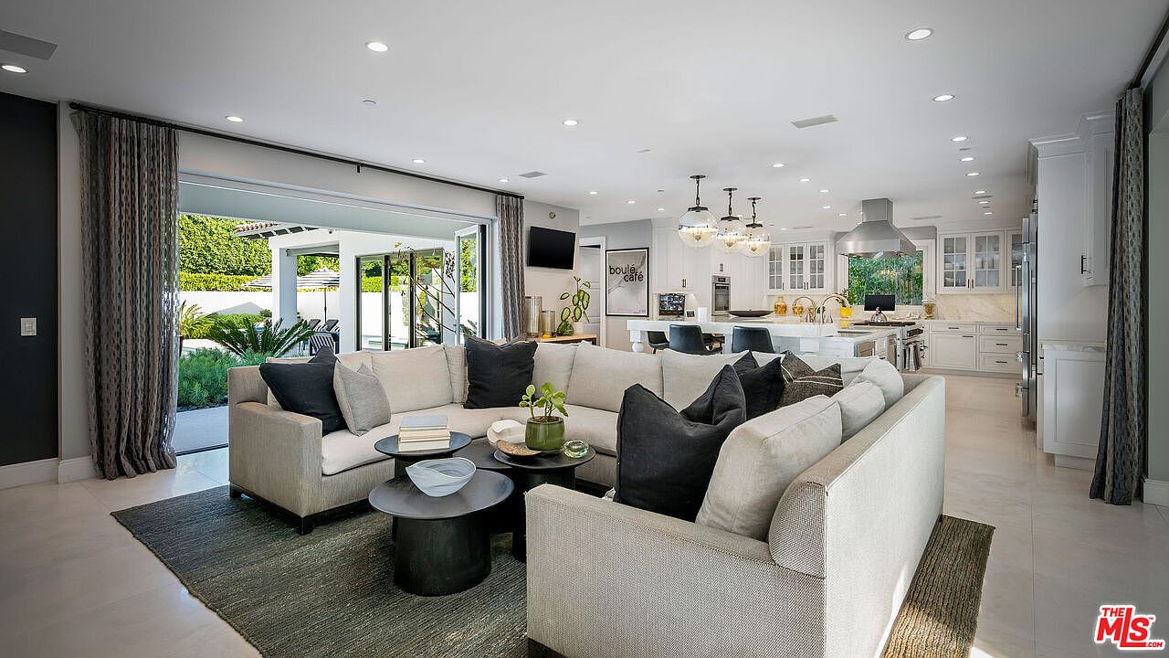 This open-concept living space features a large, neutral-toned sectional sofa arranged around a set of tiered coffee tables, set atop a textured area rug. The room seamlessly transitions to an outdoor patio through wide, retractable glass doors, creating an expansive indoor-outdoor living experience. In the background, a bright, white-toned kitchen with a central island and pendant lighting completes the airy, modern aesthetic of the home.
