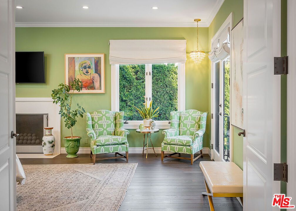 This inviting living room features vibrant green walls and a pair of patterned wingback chairs positioned near a large window that overlooks lush greenery. To the left, a fireplace with a white mantel and a decorative vase adds a classic touch, while a potted plant and a small gold-accented side table complete the seating area. The room is finished with dark wood flooring and a large area rug, creating a bright and sophisticated atmosphere.