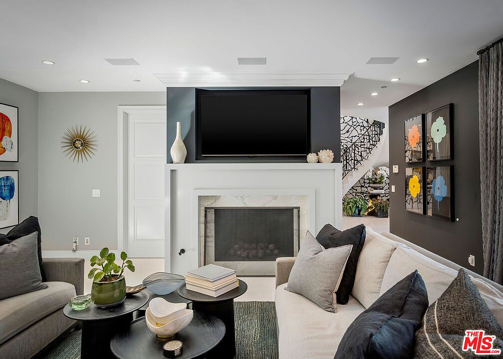 This sophisticated living room features a central fireplace with a stone surround and a mounted television above, creating a focal point for the space. The room is furnished with plush, neutral-toned sofas and layered circular coffee tables, accented by colorful floral wall art and a sunburst wall clock. A view into the background reveals a staircase with an intricate, decorative metal railing, adding a touch of architectural elegance to the open-concept layout.