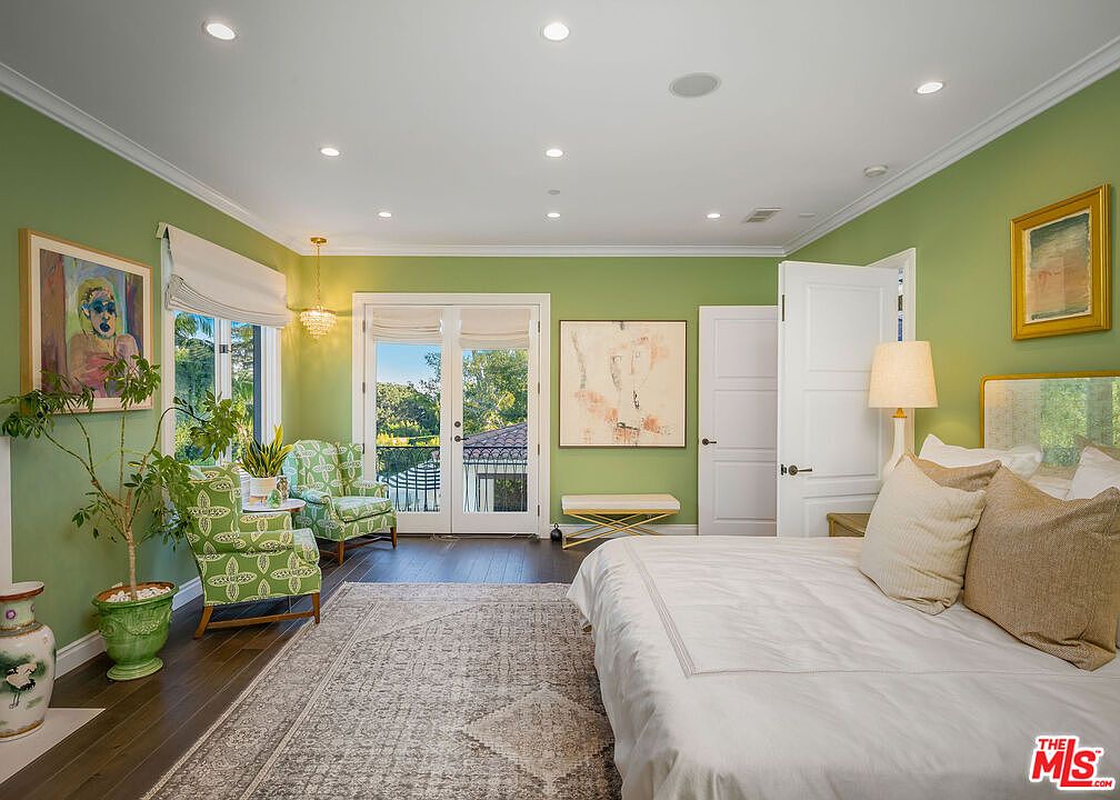 This primary bedroom features sage green walls, dark hardwood flooring, and a large area rug, creating a serene and inviting atmosphere. The room is furnished with a plush bed, two patterned armchairs near a set of French doors that open to a balcony, and decorative artwork on the walls. Recessed lighting and a crystal chandelier provide ample illumination, while the neutral bedding and light-toned furniture add a touch of elegance to the space.