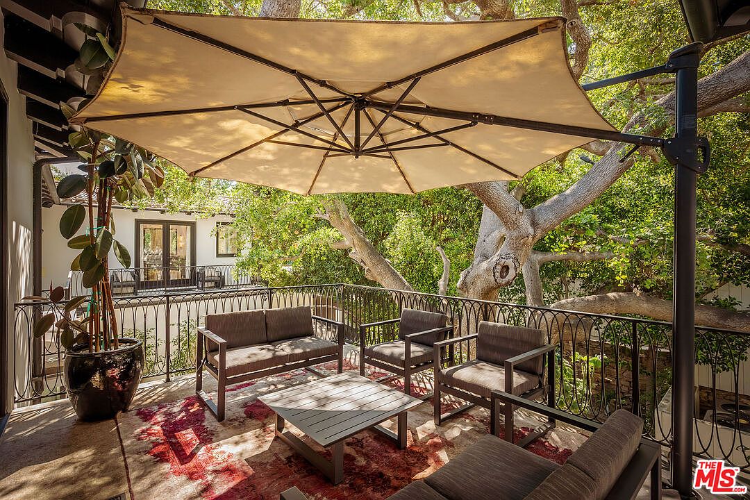 This elevated balcony features a comfortable outdoor seating area shaded by a large, cantilevered beige umbrella. The space is furnished with a sofa, two armchairs, and a central coffee table, all arranged on a patterned area rug. A decorative metal railing overlooks a lush, tree-filled backyard, creating a serene and private atmosphere.