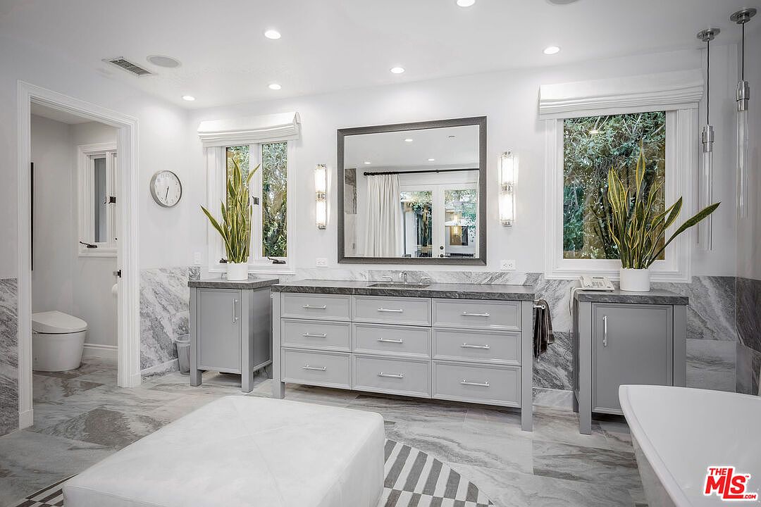 This bright and spacious primary bathroom features a double vanity with a dark stone countertop and light gray cabinetry. The room is accented by patterned stone flooring, a large central mirror, and two tall windows flanking the vanity that bring in natural light. A white ottoman sits in the foreground, and a separate water closet is visible to the left, creating a clean and sophisticated aesthetic.