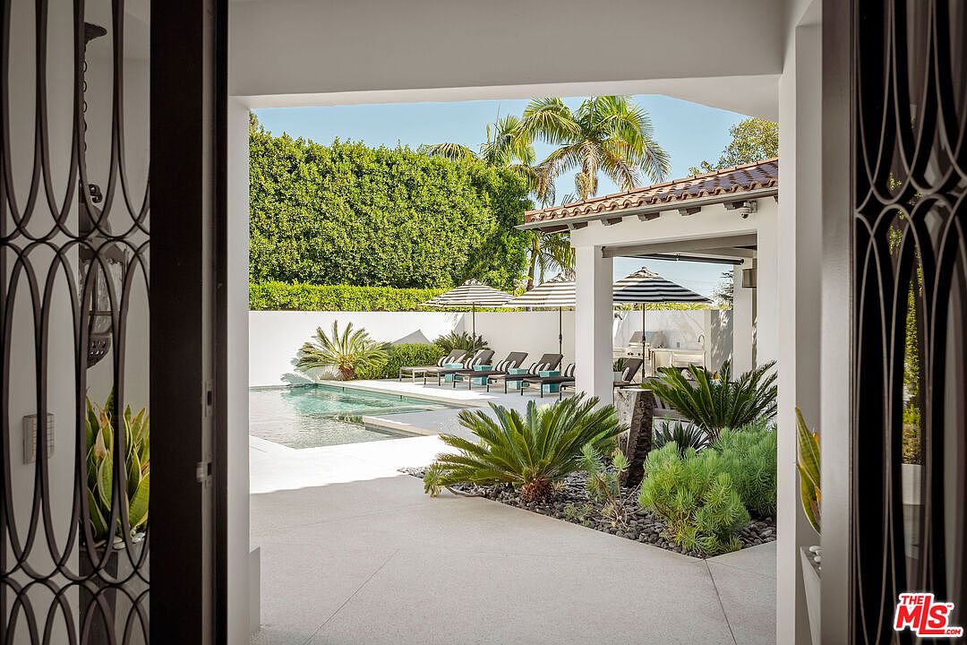 This view is captured from an interior doorway looking out toward a bright, sun-drenched backyard featuring a rectangular swimming pool. The outdoor space includes a row of lounge chairs shaded by striped umbrellas, a covered patio area with a grill, and lush landscaping with palm trees and manicured hedges. The perspective creates a seamless transition between the indoor living space and the private, resort-style outdoor retreat.