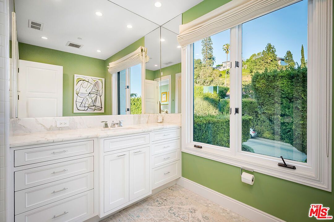 This bright and airy bathroom features a long vanity with white cabinetry and a light-colored stone countertop. A large mirror spans the wall above the vanity, reflecting the green-painted walls and a piece of abstract wall art. A large window with a fabric shade provides natural light and a view of the lush greenery outside.