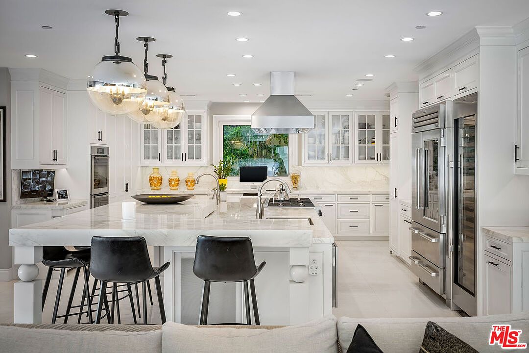 This bright, contemporary kitchen features white cabinetry, a large central island with a stone countertop, and high-end stainless steel appliances. Three glass pendant lights hang above the island, which is paired with two dark stools, while a large window above the sink provides a view of the outdoors. The space is illuminated by recessed lighting and maintains a clean, sophisticated aesthetic.