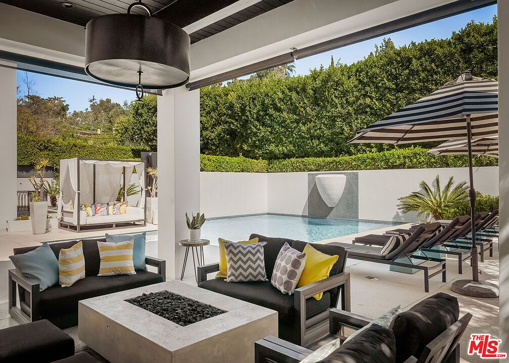 This luxurious covered patio area features a modern concrete fire pit as a central focal point, surrounded by comfortable outdoor seating with vibrant accent pillows. The space seamlessly transitions to a pristine swimming pool with a unique wall-mounted water feature, all framed by lush, manicured hedges. The overall aesthetic is sophisticated and resort-like, perfect for high-end outdoor entertaining.