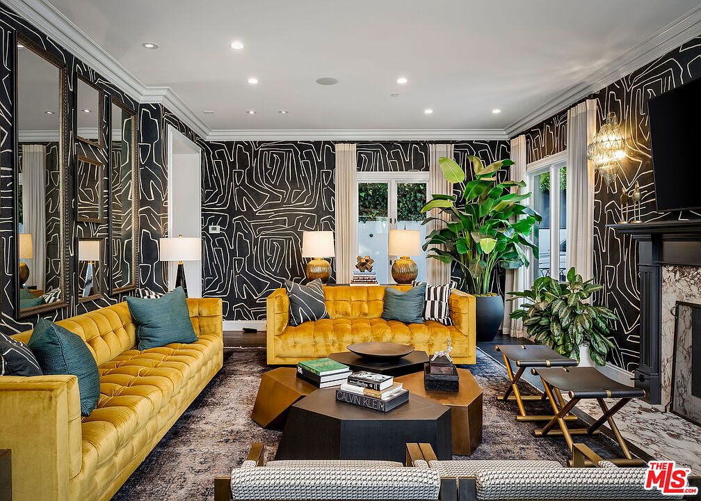 This sophisticated living room features bold, graphic black-and-white wallpaper and two vibrant mustard-yellow tufted sofas arranged around a geometric, multi-tiered coffee table. The space is accented by large potted plants, a dark patterned area rug, and a series of wall-mounted mirrors that enhance the room's depth. A fireplace with a dark surround and a mantel sits to the right, contributing to the room's dramatic and curated aesthetic.