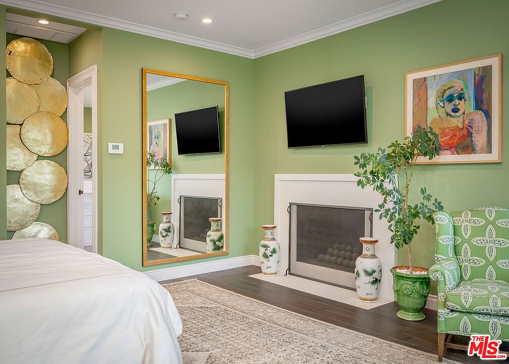 This primary bedroom features a vibrant green color palette, a large mirror with a gold frame, and a fireplace with a white surround. A patterned armchair sits beside a potted plant, while a unique wall installation of gold circular discs adds a decorative touch to the left side of the room. The space is anchored by a neutral-toned area rug over dark wood flooring, creating a sophisticated and eclectic atmosphere.
