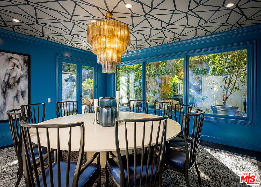 This sophisticated dining room features bold blue walls and a striking geometric patterned ceiling. A large, round white table is surrounded by dark-toned chairs, centered beneath an ornate, multi-tiered amber glass chandelier. Large windows provide a clear view of the lush greenery outside, creating a bright and elegant atmosphere.