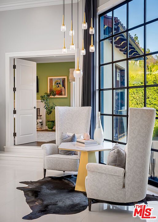This elegant seating area features two high-backed, light-colored upholstered chairs arranged around a small, gold-based table. A striking cluster of modern pendant lights hangs above, while a large, black-framed window provides a view of the exterior greenery. A dark, organic-shaped rug anchors the space, and a doorway leads into an adjacent room with green walls and a piece of artwork.