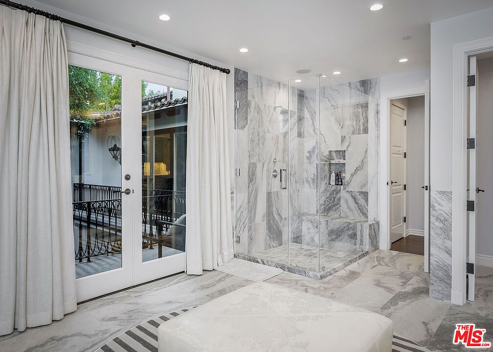 This luxurious primary bathroom features extensive use of light-toned, veined stone on both the floors and the walk-in shower walls. A glass-enclosed shower with a built-in bench sits adjacent to a set of French doors that open onto a private balcony, allowing for natural light to fill the space. The room is finished with recessed lighting and a neutral color palette, creating a bright and sophisticated atmosphere.