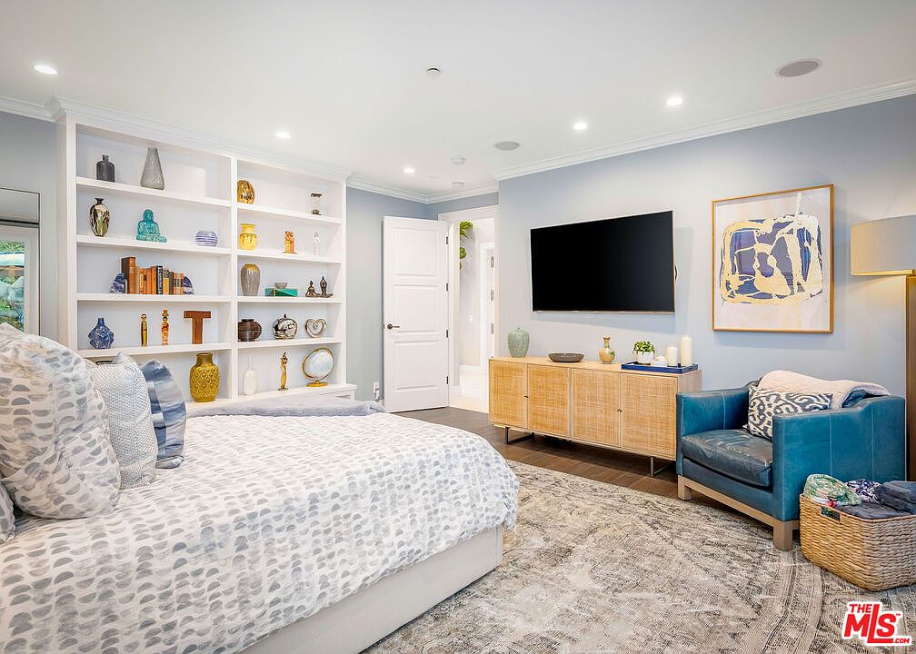 This spacious bedroom features a light blue color palette, a large bed with a patterned duvet, and a built-in shelving unit filled with decorative objects. A television is mounted on the wall above a light wood media console, complemented by a vibrant abstract painting and a comfortable blue armchair. The room is illuminated by recessed lighting and offers a serene, well-organized atmosphere.