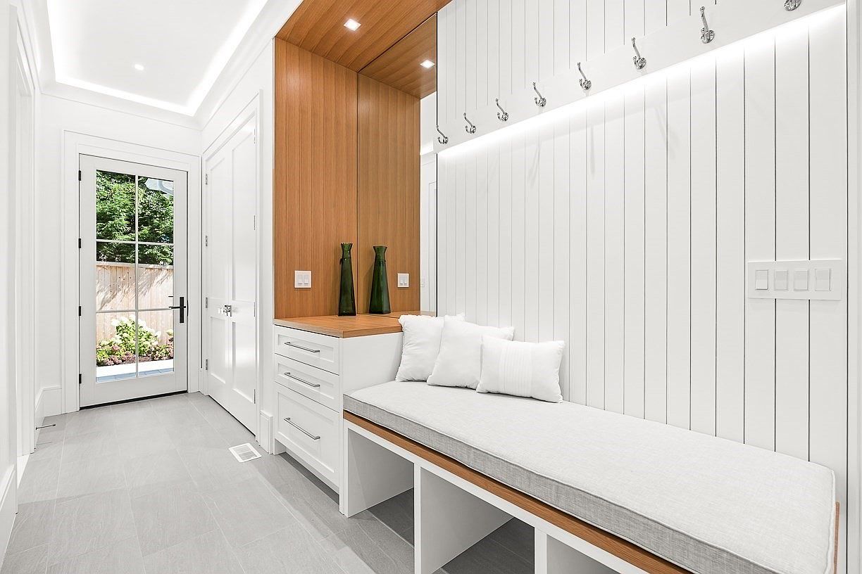 This bright and modern mudroom entryway features a built-in bench with storage cubbies, white vertical paneling, and a row of coat hooks. A light wood-toned cabinet section adds warmth to the space, complemented by a glass-paneled door that leads to an outdoor area. The clean lines and neutral color palette create a welcoming and organized transition space.