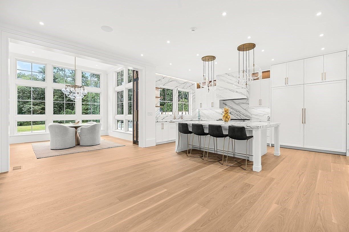 This bright, open-concept kitchen features a large central island with seating for four, complemented by white cabinetry and a striking stone backsplash. The space is illuminated by elegant pendant lighting and flows seamlessly into a dining area surrounded by large windows that offer a view of the outdoors. The light wood flooring and clean, minimalist aesthetic create a sophisticated and airy atmosphere.