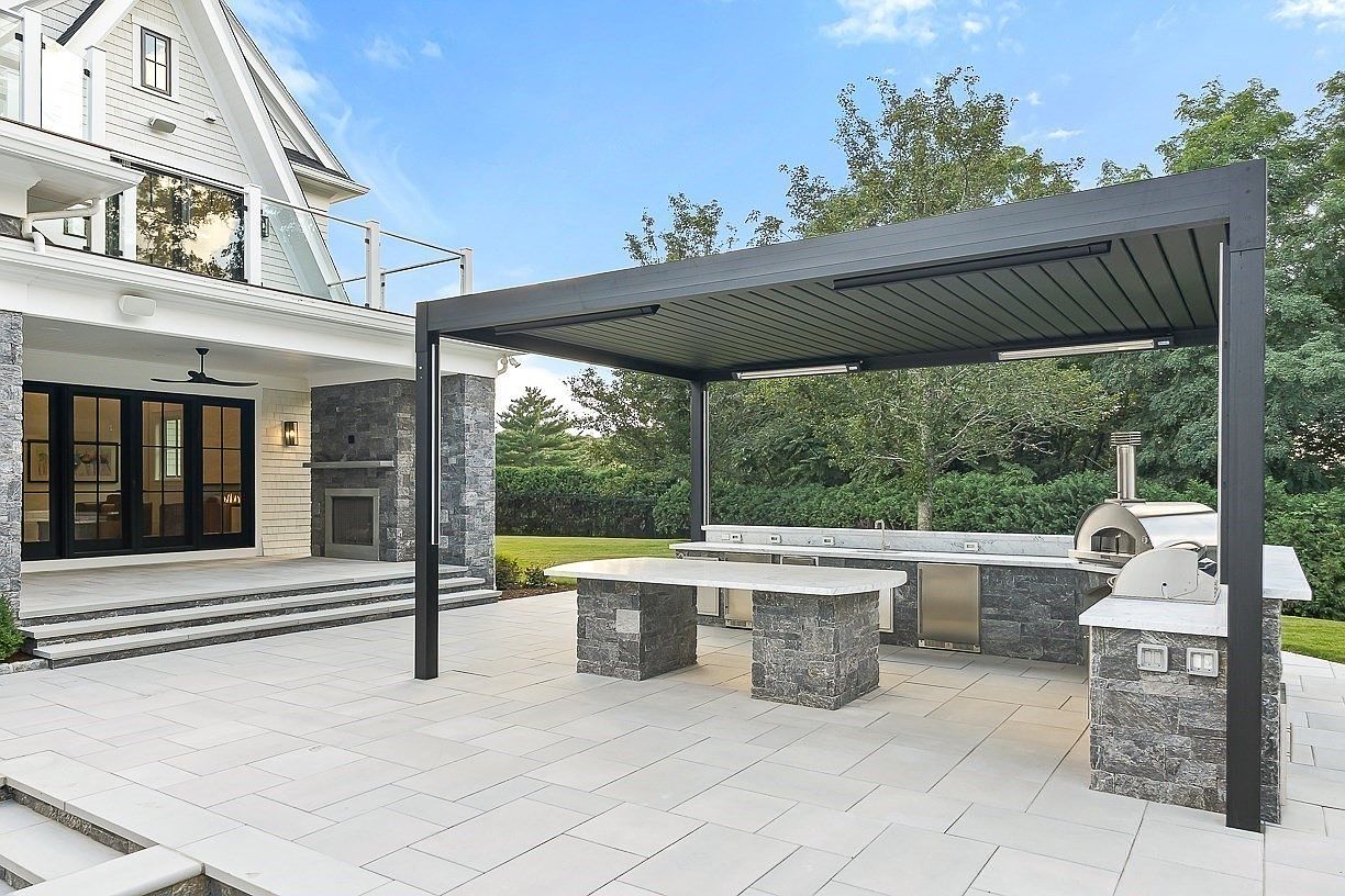 This expansive outdoor living area features a covered patio with a modern, dark-framed pergola sheltering a fully equipped outdoor kitchen. The kitchen includes a stone-clad island with a light-colored countertop, a built-in grill, and a pizza oven, all set against a backdrop of a stone-faced fireplace on the main house structure. The scene is captured from a ground-level perspective, highlighting the seamless transition between the paved patio and the lush green lawn beyond.