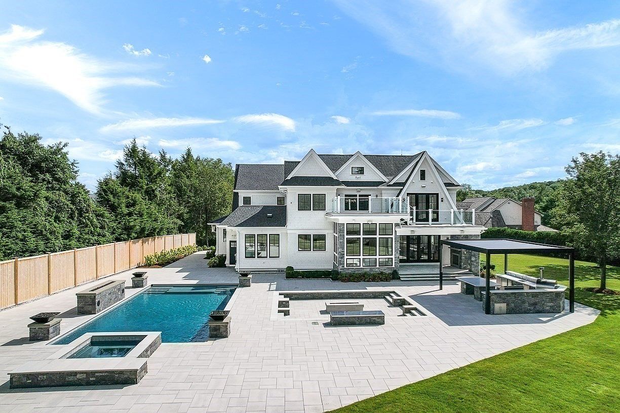 This expansive rear view of a luxury residence showcases a multi-level white shingle home with a prominent balcony and large windows. The backyard features a sophisticated hardscaped patio, a rectangular swimming pool with an integrated spa, and a covered outdoor kitchen area with a pergola. The scene is set against a backdrop of lush green trees and a clear blue sky, emphasizing a high-end, resort-style living environment.