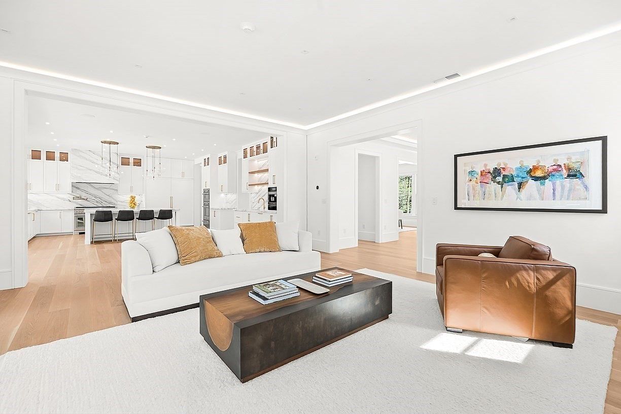This bright and airy living room features a neutral color palette with a white sofa accented by gold-toned pillows and a large, low-profile coffee table with a dark wood finish. A brown leather armchair sits on a plush white rug, while a colorful abstract painting hangs on the wall above it. The space flows seamlessly into an open-concept kitchen in the background, creating a cohesive and modern living environment.