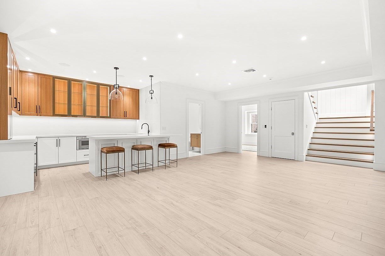 This bright, open-concept kitchen features a large central island with three bar stools and a combination of white lower cabinets and warm-toned wood upper cabinetry. The space is illuminated by recessed lighting and two pendant lights, with light-colored wood-look flooring extending throughout the room. To the right, a staircase with wooden treads leads to an upper level, creating a seamless transition between the kitchen and the rest of the home.