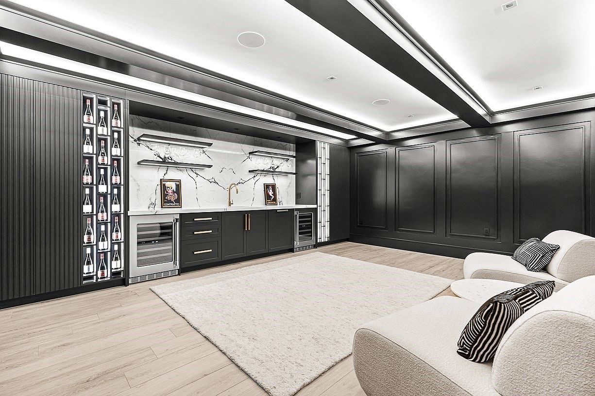 This sophisticated lounge area features a sleek wet bar with a marble-patterned backsplash, dark cabinetry, and integrated wine storage. The room is defined by its high-contrast aesthetic, combining dark paneled walls with light wood-look flooring and a plush, neutral-toned area rug. Two modern, curved white armchairs with patterned throw pillows provide a comfortable seating arrangement, creating an inviting space for entertaining.