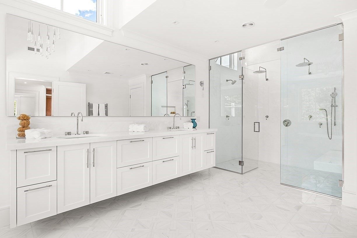 This bright and spacious primary bathroom features a long, white double vanity with ample storage and a large, wall-spanning mirror. To the right, a glass-enclosed walk-in shower with multiple showerheads offers a clean, modern aesthetic. The room is finished with light-toned patterned tile flooring, creating a cohesive and luxurious atmosphere.
