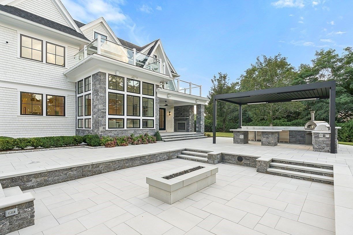 This expansive stone patio features a sunken fire pit area and a covered outdoor kitchen equipped with a built-in grill and pizza oven. The rear of the house showcases a sophisticated blend of white siding and stone accents, with large windows and a second-story balcony overlooking the space. The scene is captured from a ground-level perspective, emphasizing the seamless transition between the indoor living areas and the luxurious outdoor entertainment zone.