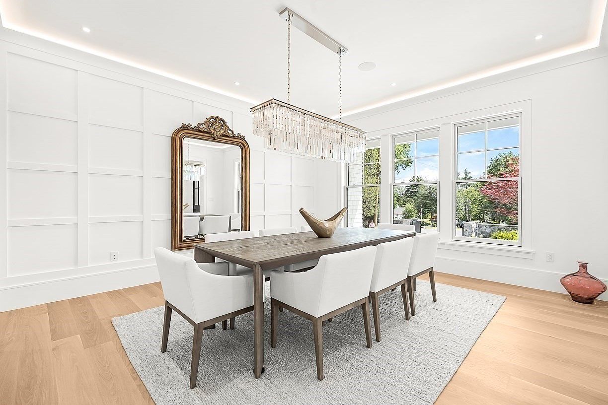 This elegant dining room features a long wooden table surrounded by six upholstered chairs, set atop a light-colored area rug. A large, ornate gold-framed mirror leans against a white paneled wall, while a crystal chandelier hangs above the table as a focal point. Large windows allow natural light to fill the space, highlighting the light wood flooring and the clean, sophisticated aesthetic of the room.