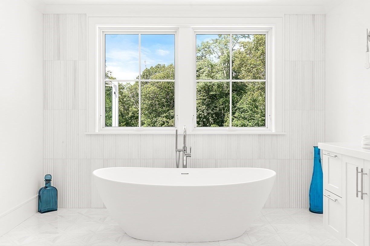 This bright and airy primary bathroom features a sleek, freestanding soaking tub centered beneath a pair of large windows that overlook lush greenery. The walls are adorned with vertical textured tile, creating a clean, modern aesthetic, while a white vanity with a stone countertop sits to the right. Two vibrant blue glass vases provide a striking pop of color against the neutral, light-filled space.