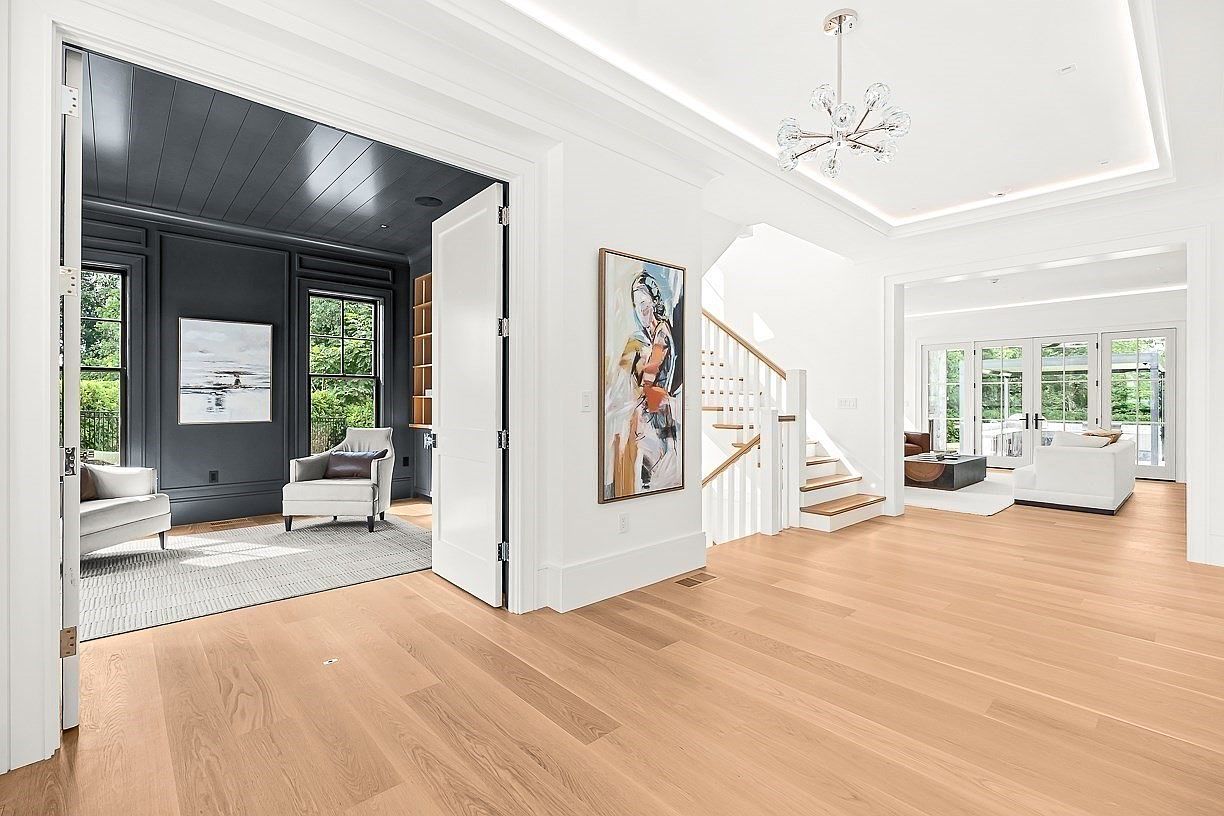This bright and airy foyer features light wood flooring and a central staircase with white railings. To the left, a dark-walled office space provides a striking contrast, while the open-concept living area to the right is filled with natural light from large windows. A modern, multi-armed chandelier hangs from the tray ceiling, adding a touch of elegance to the clean, contemporary design.