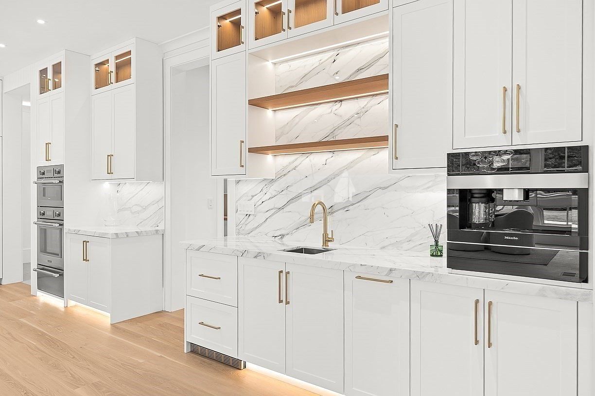 This bright, modern kitchen features crisp white cabinetry, light wood flooring, and a striking stone backsplash with dramatic veining. The space is equipped with high-end integrated appliances, including a built-in coffee system, and is accented by warm under-cabinet lighting and gold-toned hardware. The perspective captures a clean, functional layout with open shelving and a sleek sink area, creating an elegant and sophisticated atmosphere.