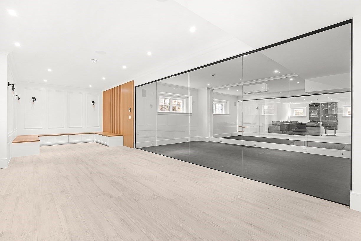 This bright, modern home gym features light wood-look flooring and a large wall of floor-to-ceiling mirrors that reflect the adjacent living space. The room includes built-in bench seating with storage underneath, accented by a vertical wood-slat feature wall. The space is illuminated by recessed lighting, creating a clean, minimalist aesthetic suitable for a workout area.