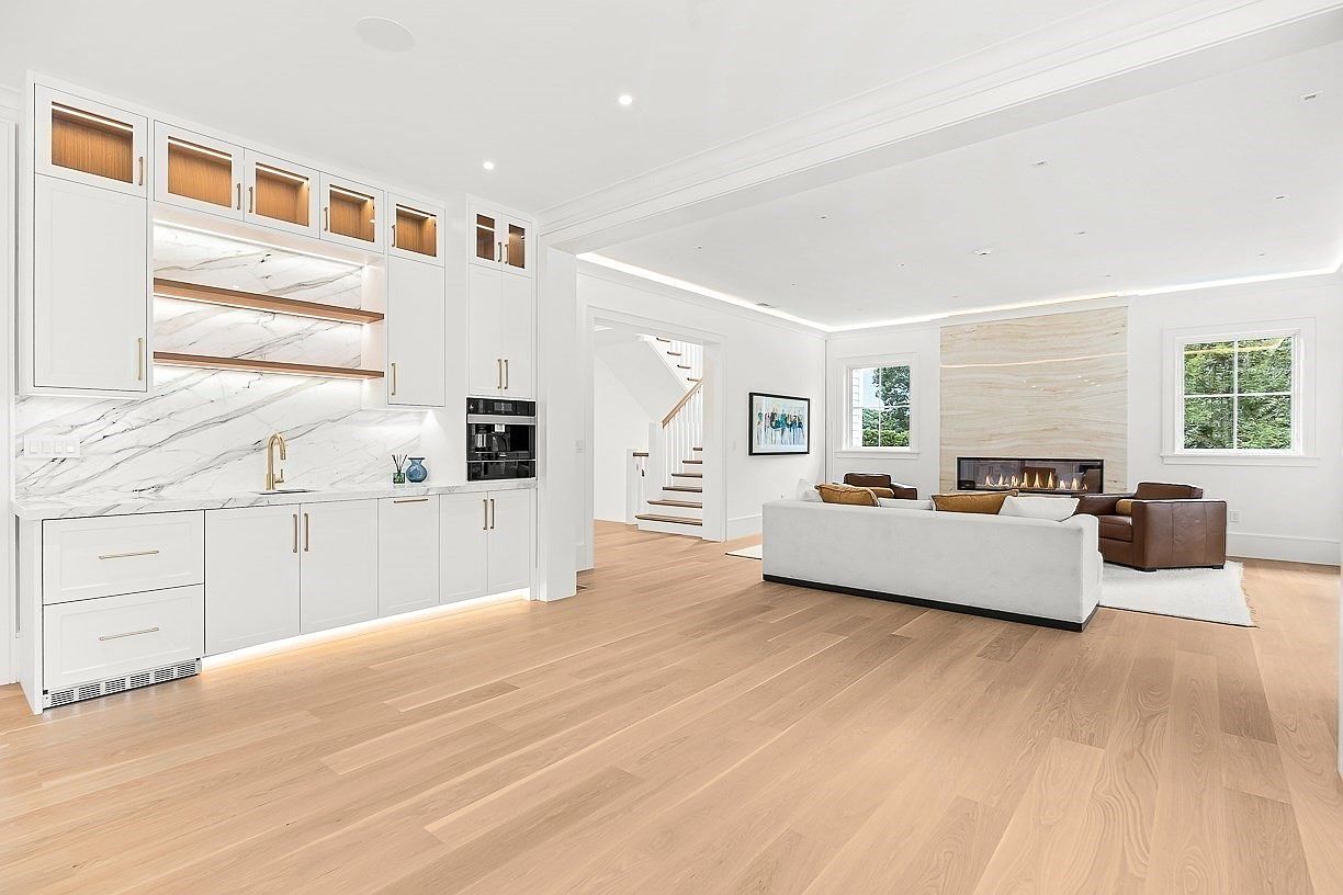 This open-concept living space features light wood flooring and a bright, neutral color palette. To the left, a built-in wet bar with white cabinetry and a stone backsplash provides a sophisticated entertaining area, while the main living area centers around a fireplace with a tall, light-toned stone surround. The room is furnished with a plush white sofa and leather armchairs, creating a comfortable and airy atmosphere that flows toward a staircase in the background.